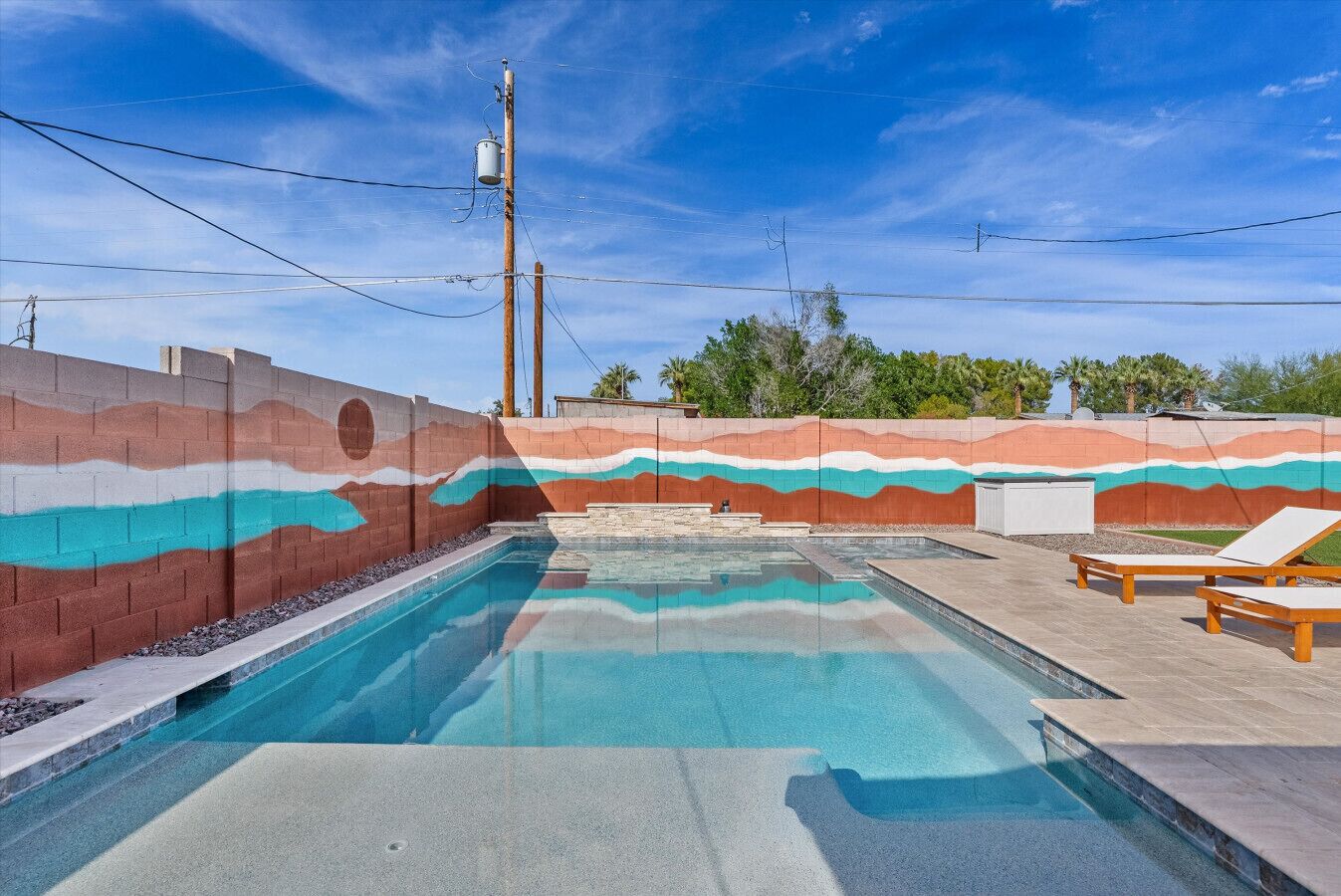 Ein Swimmingpool mit farbenfrohem Wandgemälde, einer Holzterrasse und Liegestühlen.