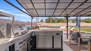 Een buitenkeuken met een Char-Broil grill, een spoelbak en een inbouwkoelkast. Er zijn loungestoelen en een overdekte patio.