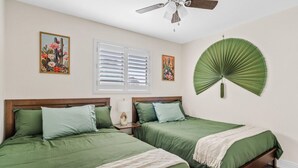 Een slaapkamer met twee bedden, een plafondventilator, een raam met zonwering en een ventilator aan de muur.