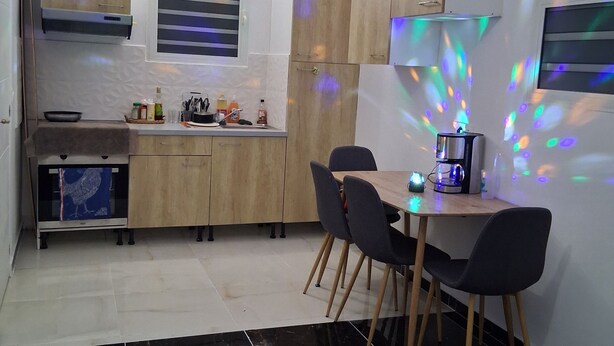 A modern kitchen with wooden cabinets, a dining area with black chairs, and a black-tiled floor. - Espace Zen Bellevue (Sainte-Rose)