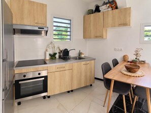A modern kitchen with a built-in oven, a stainless steel range hood, and a dining area with a wooden table and chairs. - Ti Vétiver (Sainte-Rose)