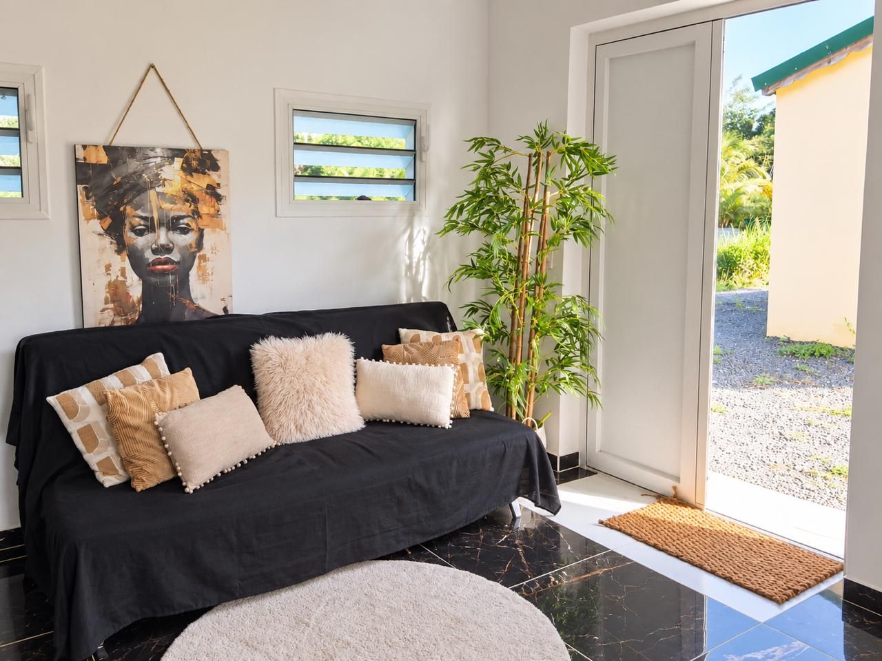 Una sala moderna con un sofá negro, almohadones decorativos, un cuadro grande, una planta de bambú y vistas a través de un ventanal.