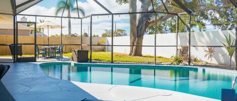 Una piscina al aire libre techada con sillas de descanso y una mesa, rodeada por una valla de madera y árboles.