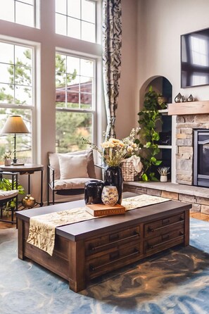 A cozy living room with a large window, a fireplace, a coffee table with decorative items, and a comfortable seating area. - Lakefront Spa Luxe • Theater & Game & View • USAFA 10 Min • Sleeps 10 (Monument)