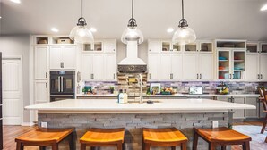 A kitchen with white cabinets, a central island with stools, a built-in oven, and pendant lighting. - Lakefront Spa Luxe • Theater & Game & View • USAFA 10 Min • Sleeps 10 (Monument)