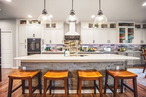 Une cuisine avec des meubles blancs, un îlot central doté de tabourets, un four encastré et des luminaires suspendus.