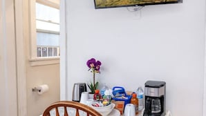Ein Essbereich mit Tisch für zwei Personen, einem Stuhl, einem an der Wand befestigten Fernseher, der eine Landschaft zeigt, und einem Fenster mit Blick auf einen Balkon.