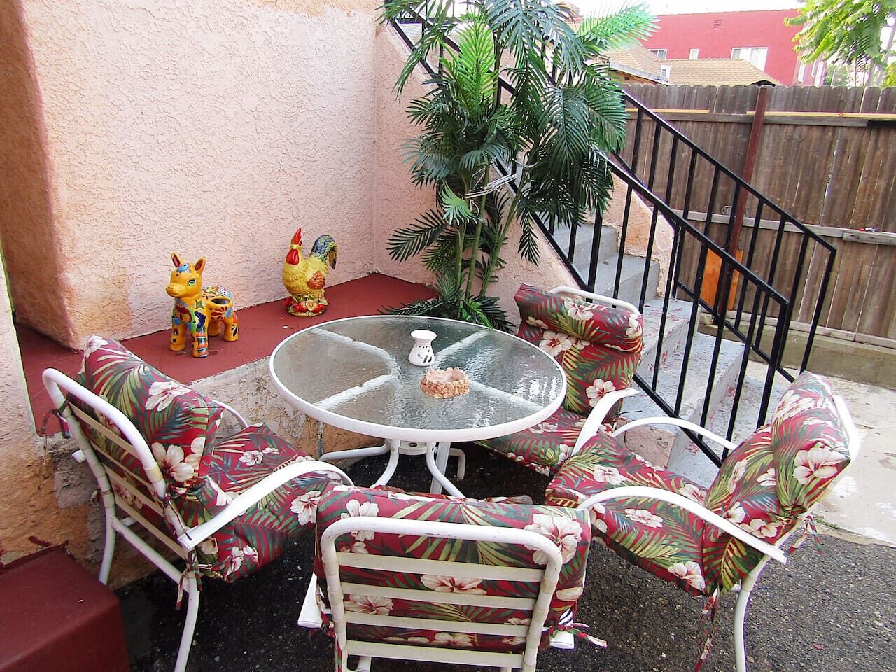 Een terras met een ronde glazen tafel en stoelen met een bloemmotief, omgeven door een houten schutting en een potplant.