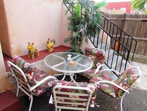 Eine Terrasse mit rundem Glastisch und stuhlförmigen Sitzgelegenheiten mit Blumenmuster, umgeben von einem Holzzaun und einem Blumentopf.