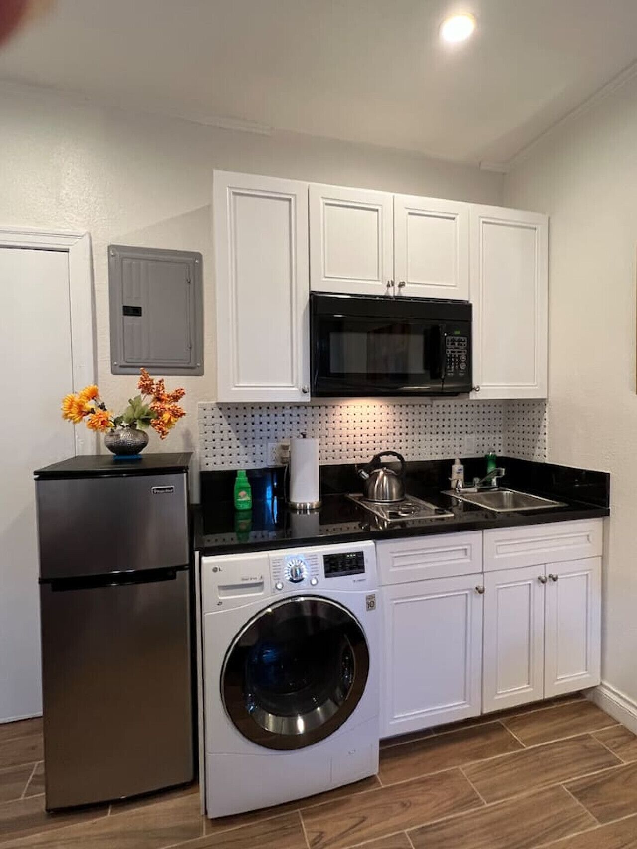 A compact kitchen with white cabinets, a stainless steel refrigerator, a black microwave, a built-in washing machine, and a sink.