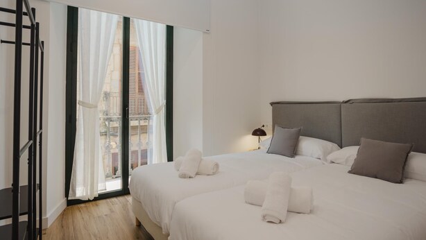 Hotelkamer met een groot bed, nachtkastje, lamp en uitzicht op een gebouw door een raam met dunne gordijnen.