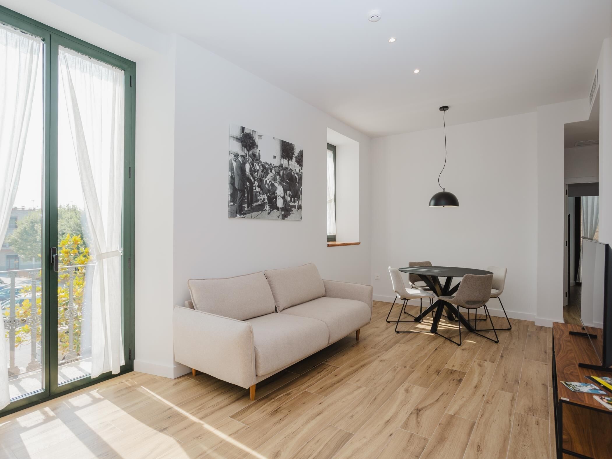 Una sala de estar moderna con sofá, mesa de comedor y sillas. Hay un ventanal con cortinas, una lámpara colgante negra y una fotografía blanco y negro en la pared.