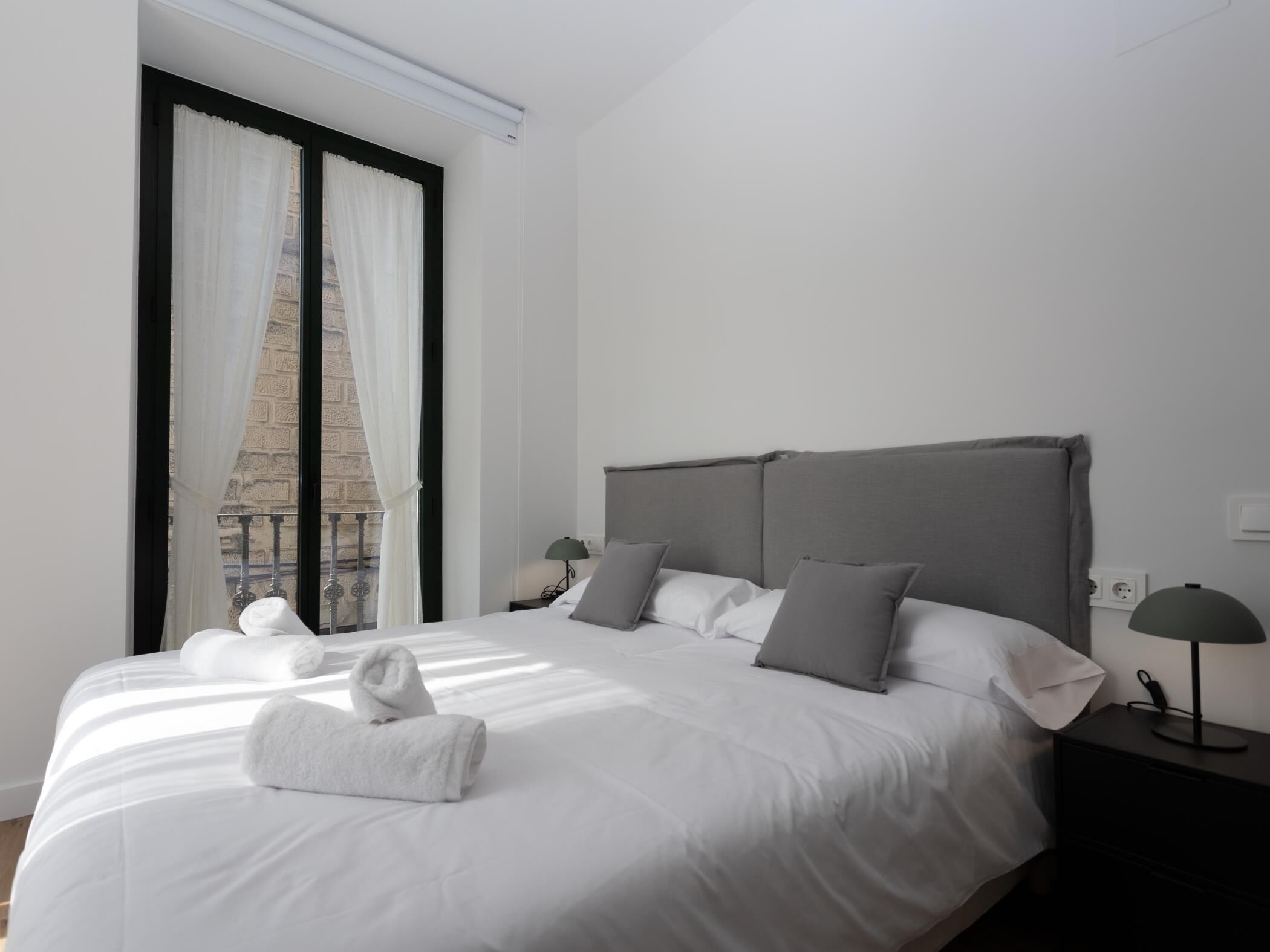 A modern hotel room with a large bed, grey headboard, and white linens. There are two bedside tables with lamps and a view of a brick building through the window.