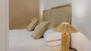 Een bed met beige beddengoed en kussens, een houten lamp en een structuurmuur.