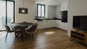 Een moderne keuken met witte kasten, een zwart aanrecht en een inbouwoven. Er is een eethoek met een ronde tafel en stoelen, en een televisie op een houten standaard.