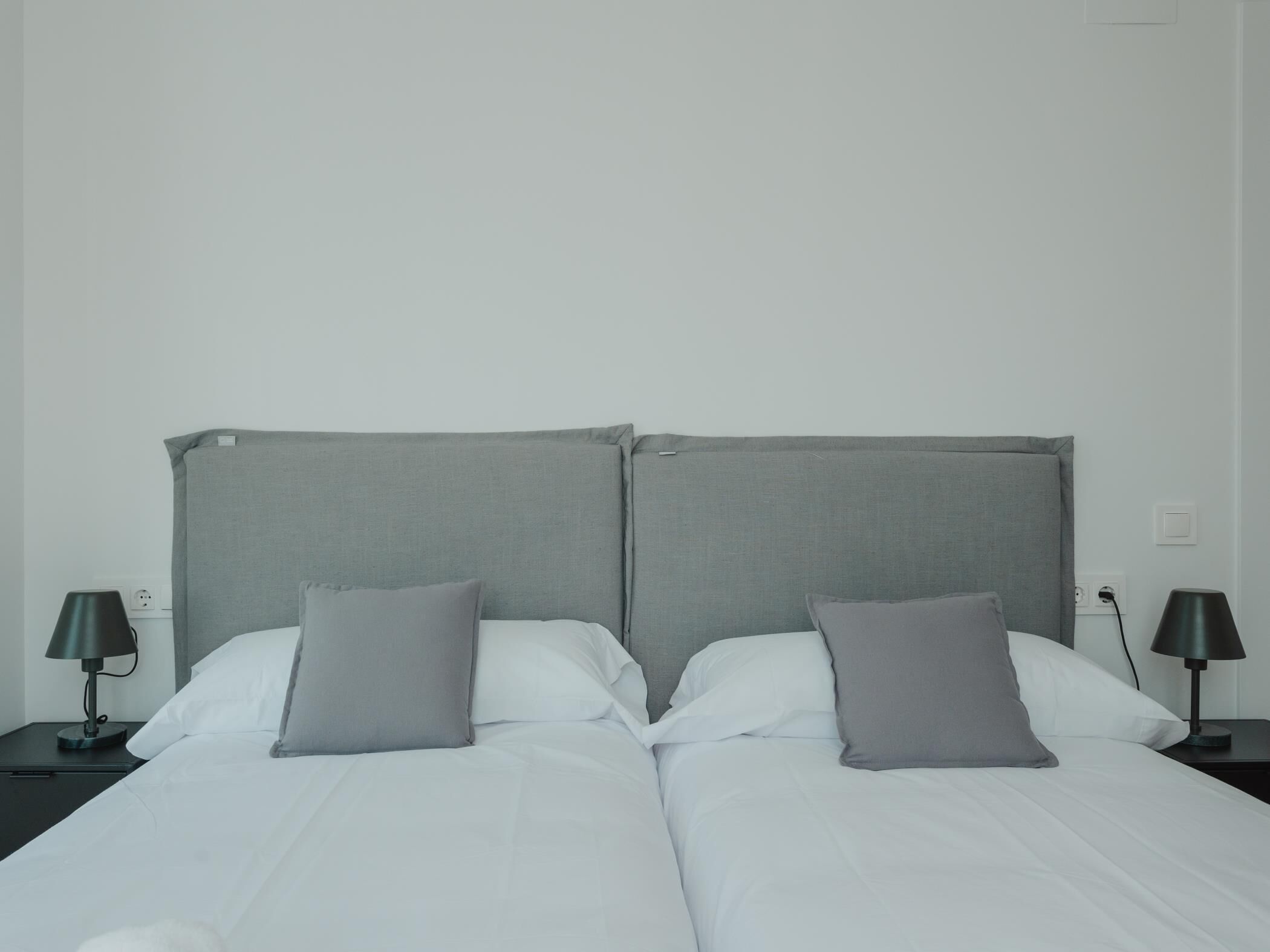 Een tweepersoonsbed met grijs hoofdbord en kussens, twee nachtkastjes met lampen, en een effen witte muur.