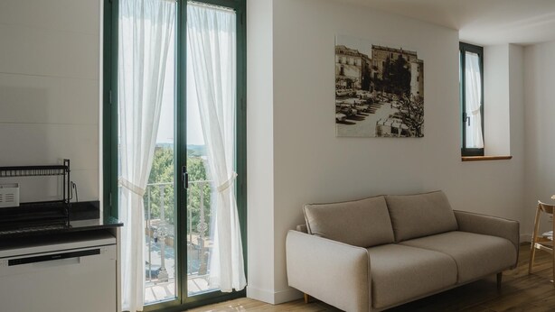Un salon moderne avec un canapé beige, un coin repas doté d’un plan de travail noir et une grande fenêtre ornée de rideaux légers.