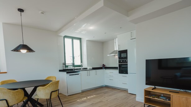 Une cuisine moderne avec des armoires blanches, un comptoir noir et un four encastré. On y trouve un coin repas avec des chaises jaunes et une table noire. Un téléviseur est fixé au mur.
