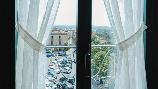 A view through a window with sheer curtains, revealing a parking lot and buildings in the distance.