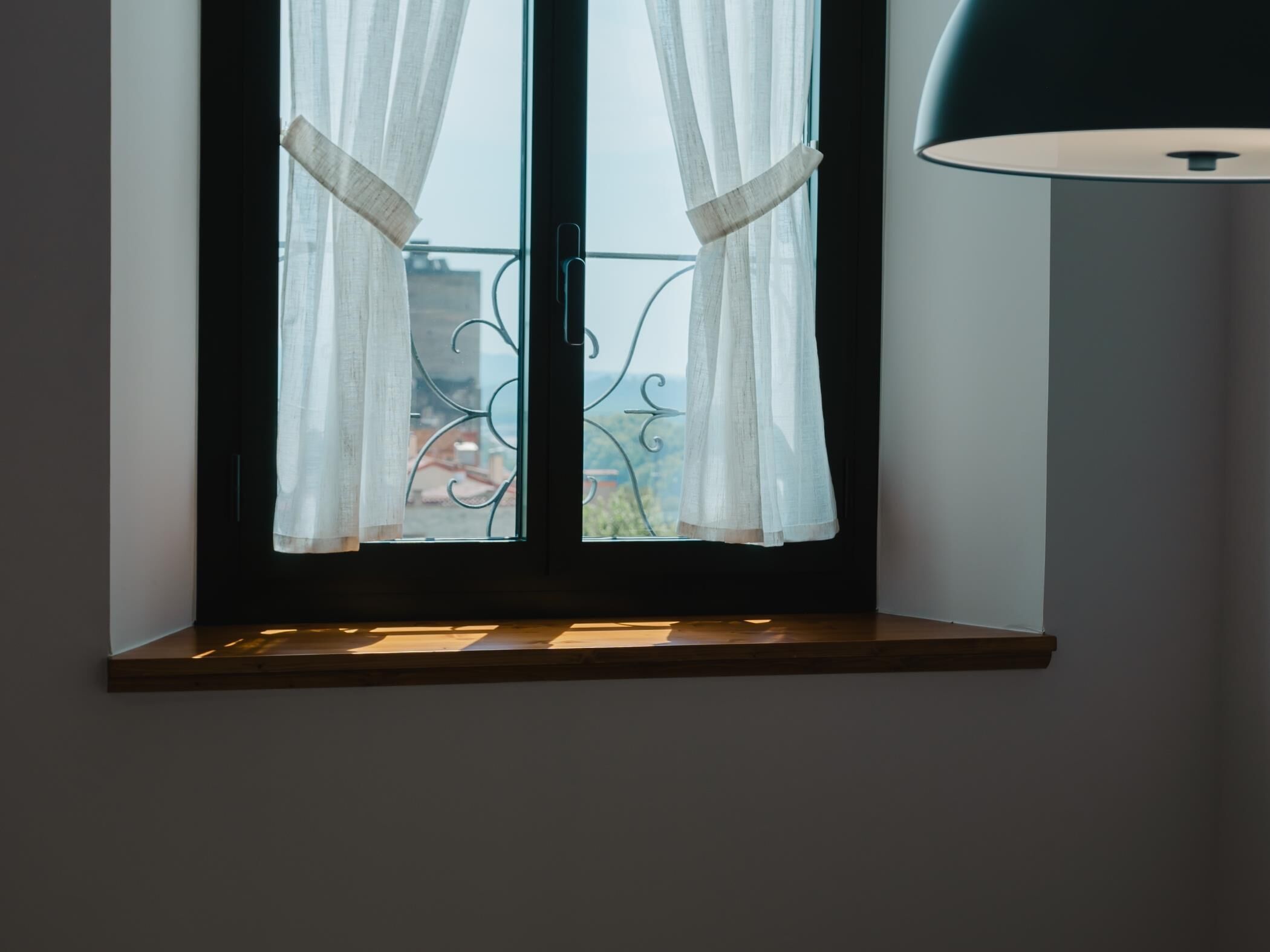 Une fenêtre avec des rideaux blancs, une rambarde de balcon en métal et une lampe noire.
