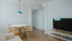 A modern living room with a white sofa, wooden dining table, and chairs, a TV on a wooden stand, and a ceiling light.