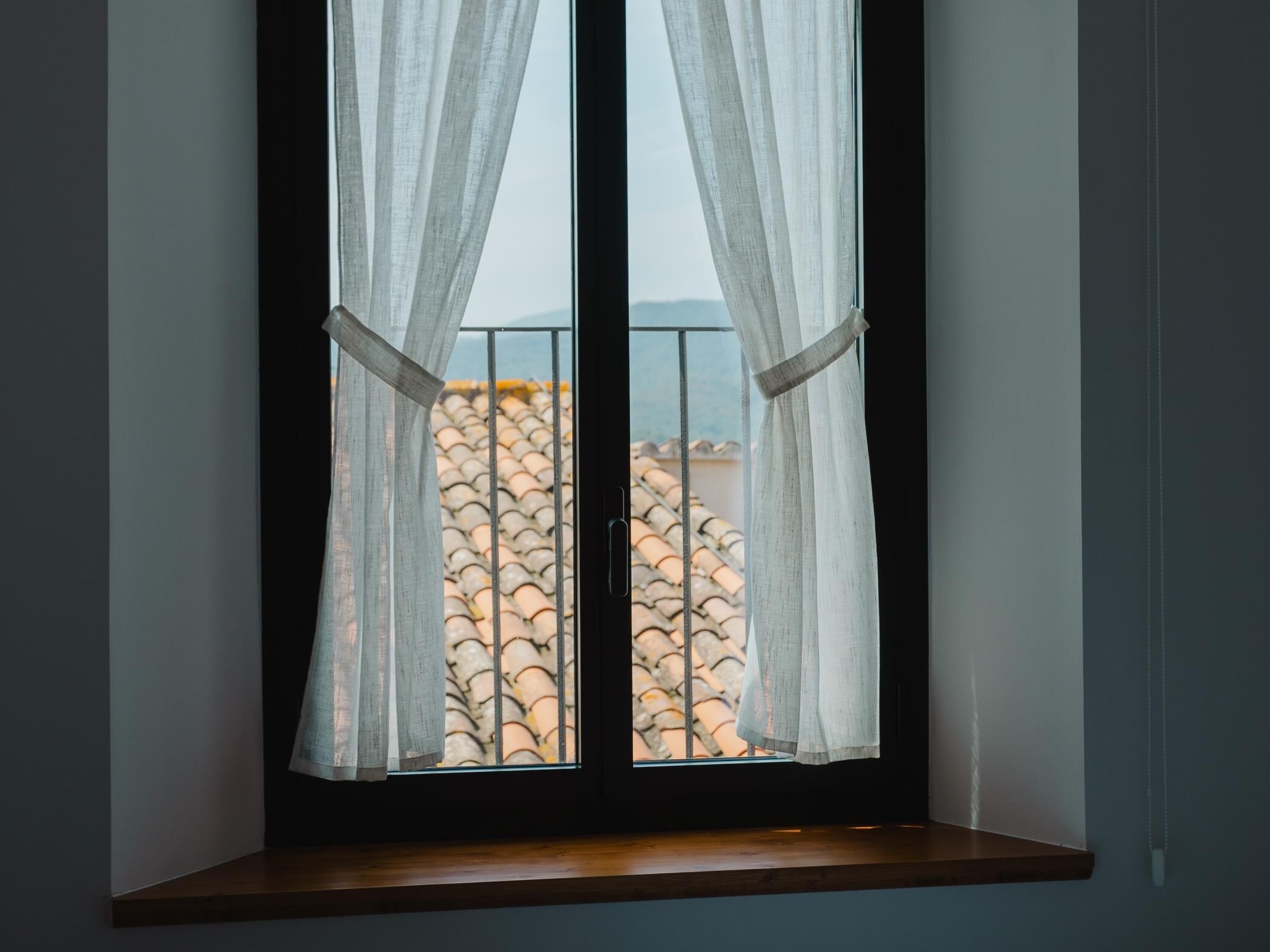 A window with white curtains, a view of a tiled roof, and a wooden ledge below.