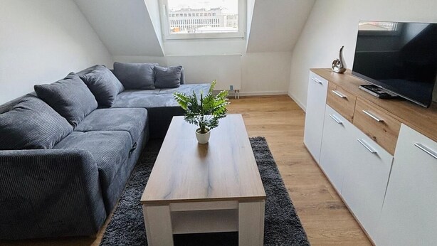 Un dormitorio estilo loft moderno con techo inclinado, un ventanal grande, un sofá gris oscuro, una mesa de centro de madera y un televisor sobre un mueble de madera.