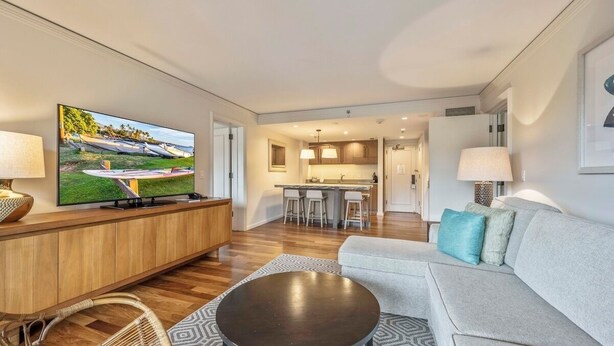 A modern living room with a flat-screen TV, a grey sofa, a round coffee table, and a kitchen area in the background. - Tropical Tranquility - Stylish Condo w/ Ocean View (Lahaina)