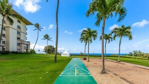 A shuffleboard court with a green surface, surrounded by palm trees and a building with multiple floors. - Tropical Tranquility - Stylish Condo w/ Ocean View (Lahaina)