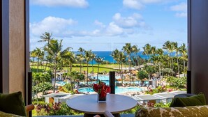 A balcony with a view of a pool and palm trees, a table with a vase of flowers, and a view of the ocean. - Tropical Tranquility - Stylish Condo w/ Ocean View (Lahaina)