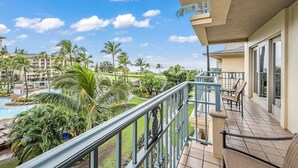 Un balcon donnant sur une piscine et des palmiers, un coin repas avec des chaises, et un bâtiment avec une enseigne.
