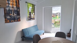 Quarto com um sofá azul, uma mesa de madeira redonda com um prato e vista para um jardim através de uma porta deslizante.