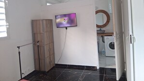 Quarto com chão em azulejos, guarda-roupa de madeira, televisão na parede, máquina de lavar roupa e um espelho.