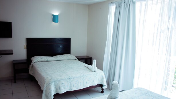 Una habitación de hotel con cama, mesita de noche, televisor en la pared y ventana con cortinas.