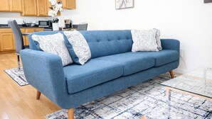 Un salon moderne avec un canapé bleu, des coussins à motifs, une table basse en verre et une cuisine en arrière-plan.