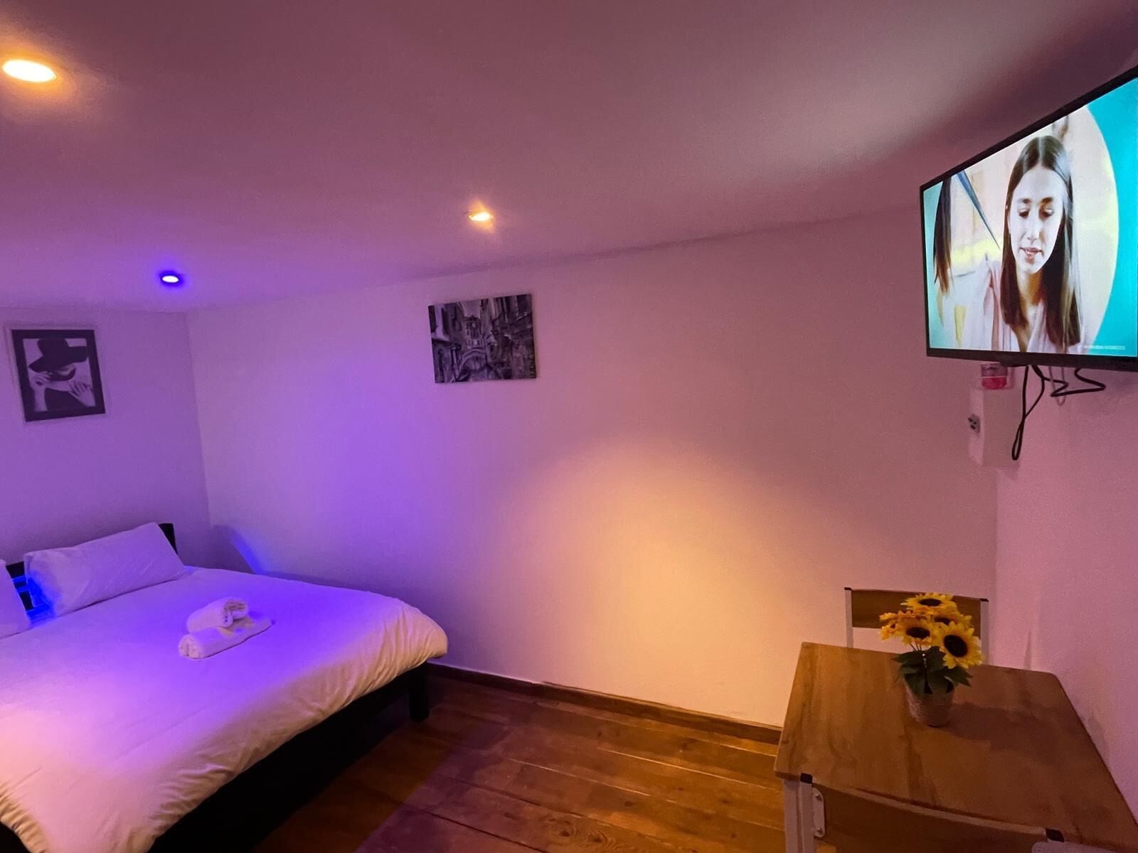 Un dormitorio con una cama, un televisor montado en la pared y una pequeña mesa de madera con un jarrón de girasoles.