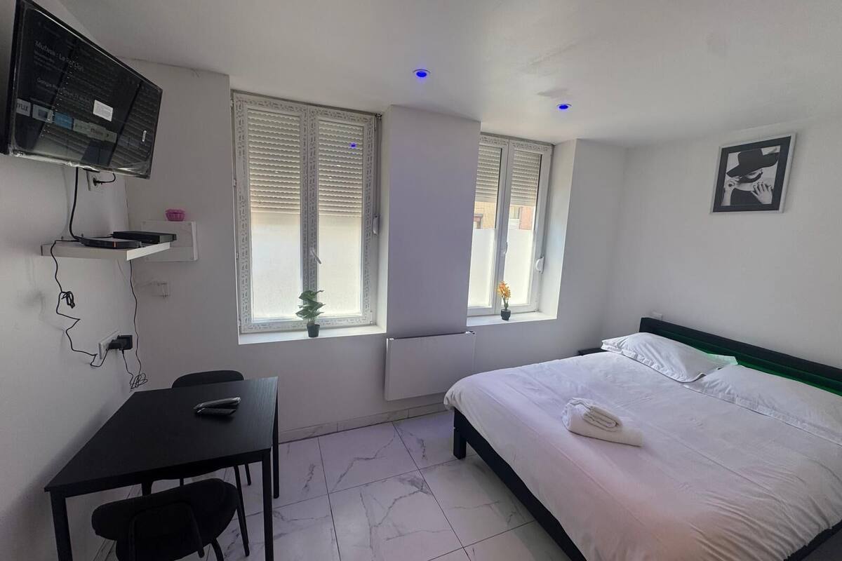 Ein Schlafzimmer mit einem Bett, einem Schreibtisch, einem Sessel, einem an der Wand montierten Fernseher und zwei Fenstern mit Jalousien.