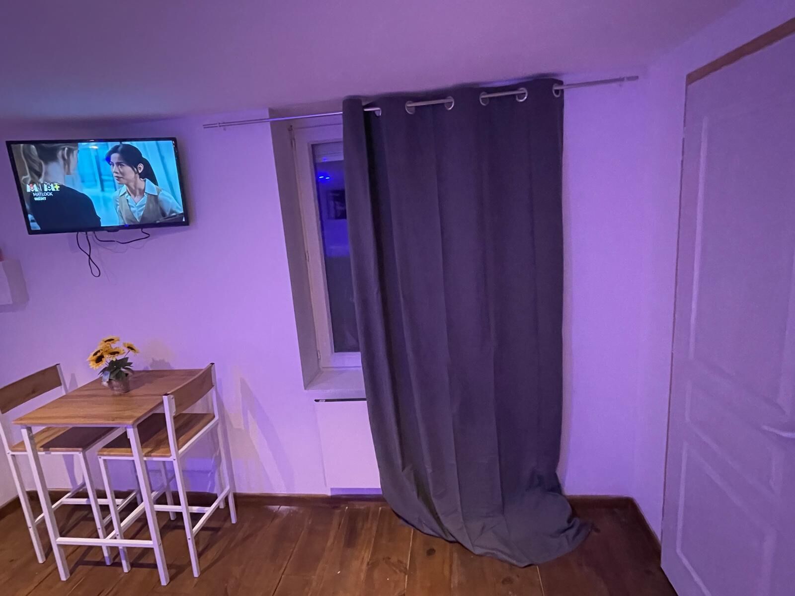 Une pièce avec une table en bois, un téléviseur fixé au mur et une fenêtre avec un rideau.