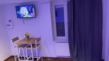 A small room with a wooden table and chairs, a TV mounted on the wall, a window with a curtain, and a door.