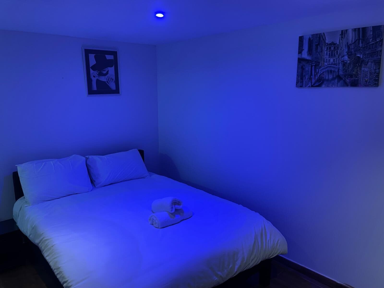 A single bed with white bedding and a rolled towel, a framed picture on the wall, and a cityscape painting.