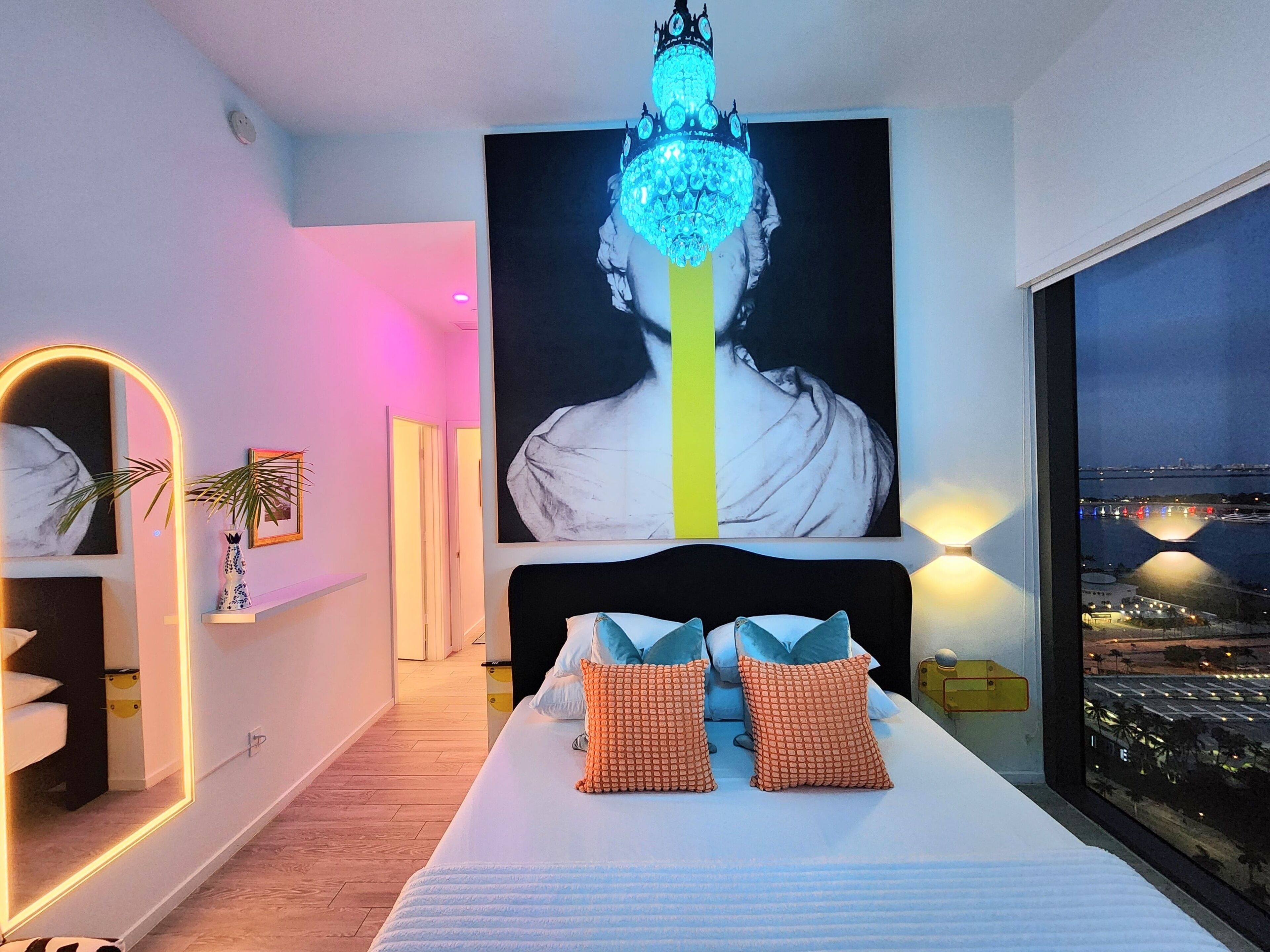 A modern bedroom with a large bed, a chandelier, a city view, and a large portrait on the wall.