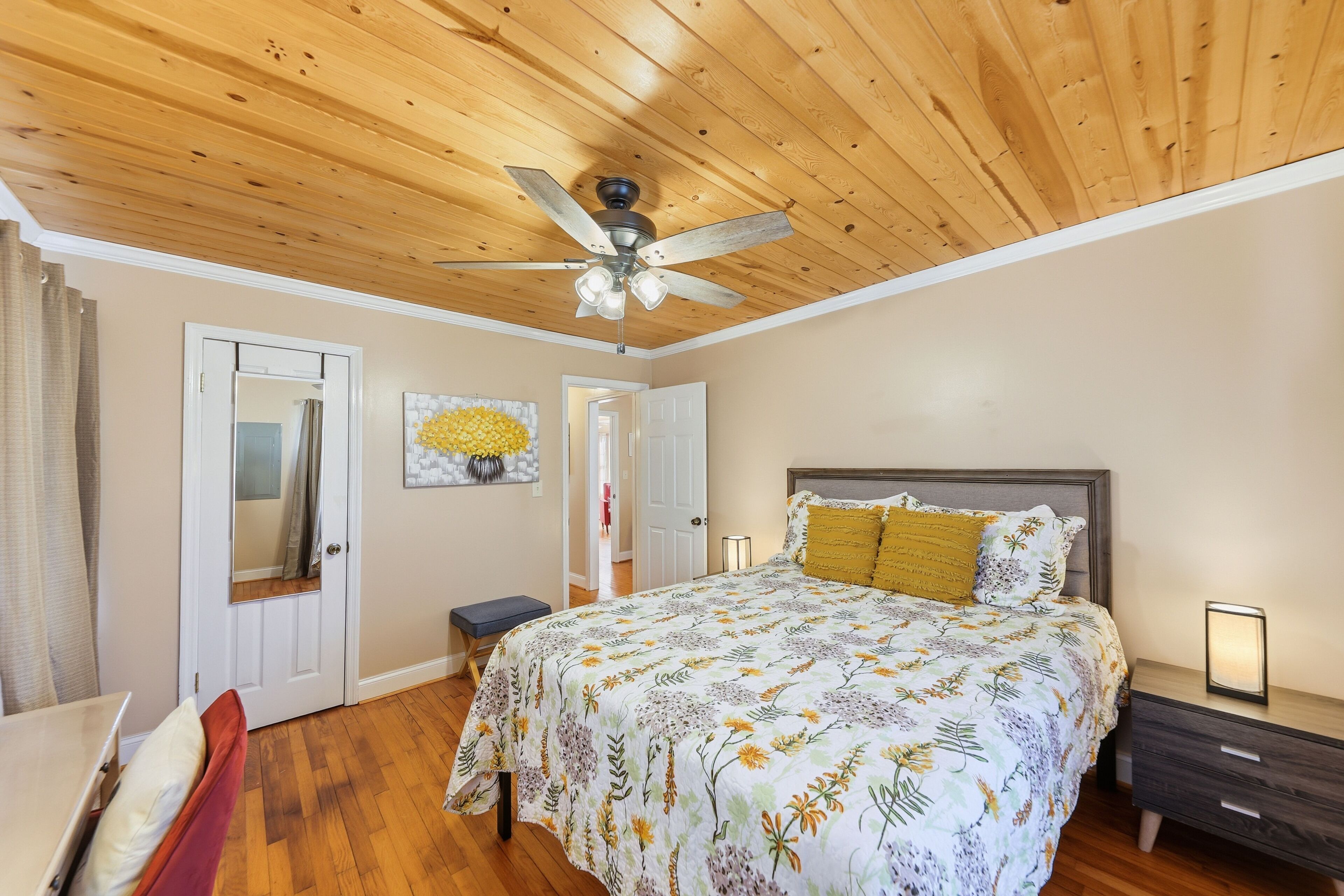 A bedroom with a wooden ceiling, a bed with a floral bedspread, a nightstand, a chair, and a door.