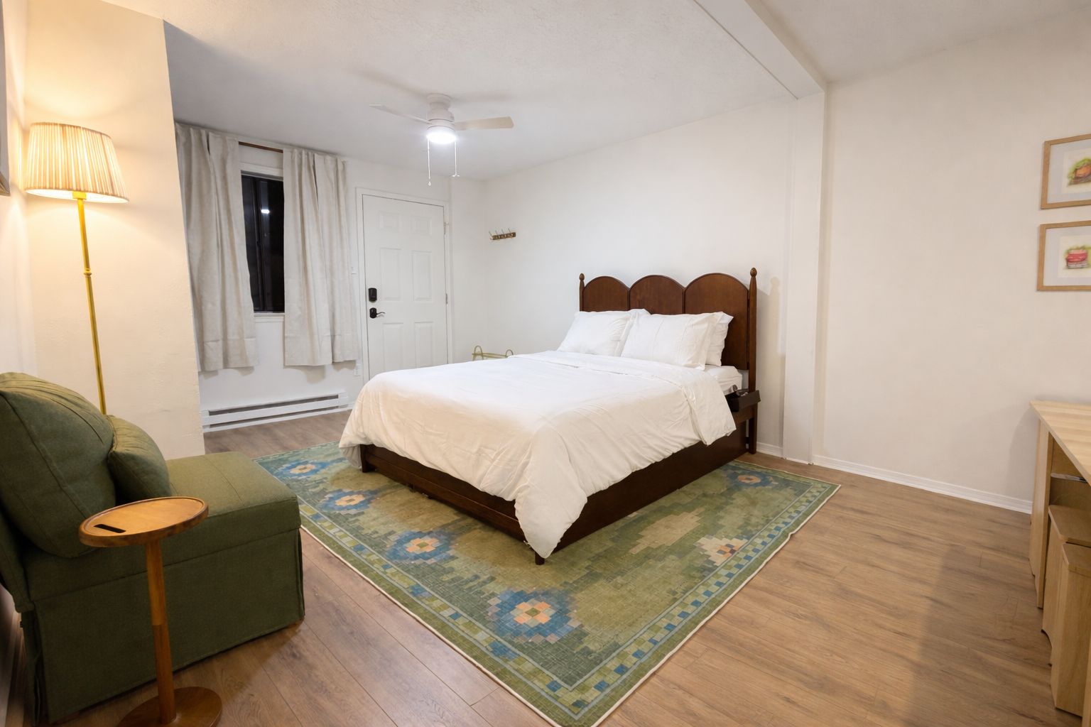 A bedroom with a wooden bed, a green armchair, a small side table, a patterned rug, and a ceiling fan.