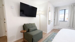 Une chambre d’hôtel avec un lit, un canapé vert, un téléviseur fixé au mur, une lampe sur pied et une fenêtre avec des rideaux.