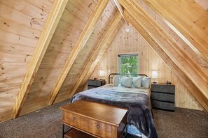 A bedroom with a sloped ceiling, a bed with pillows, a nightstand, a dresser, and a window with a view of greenery.