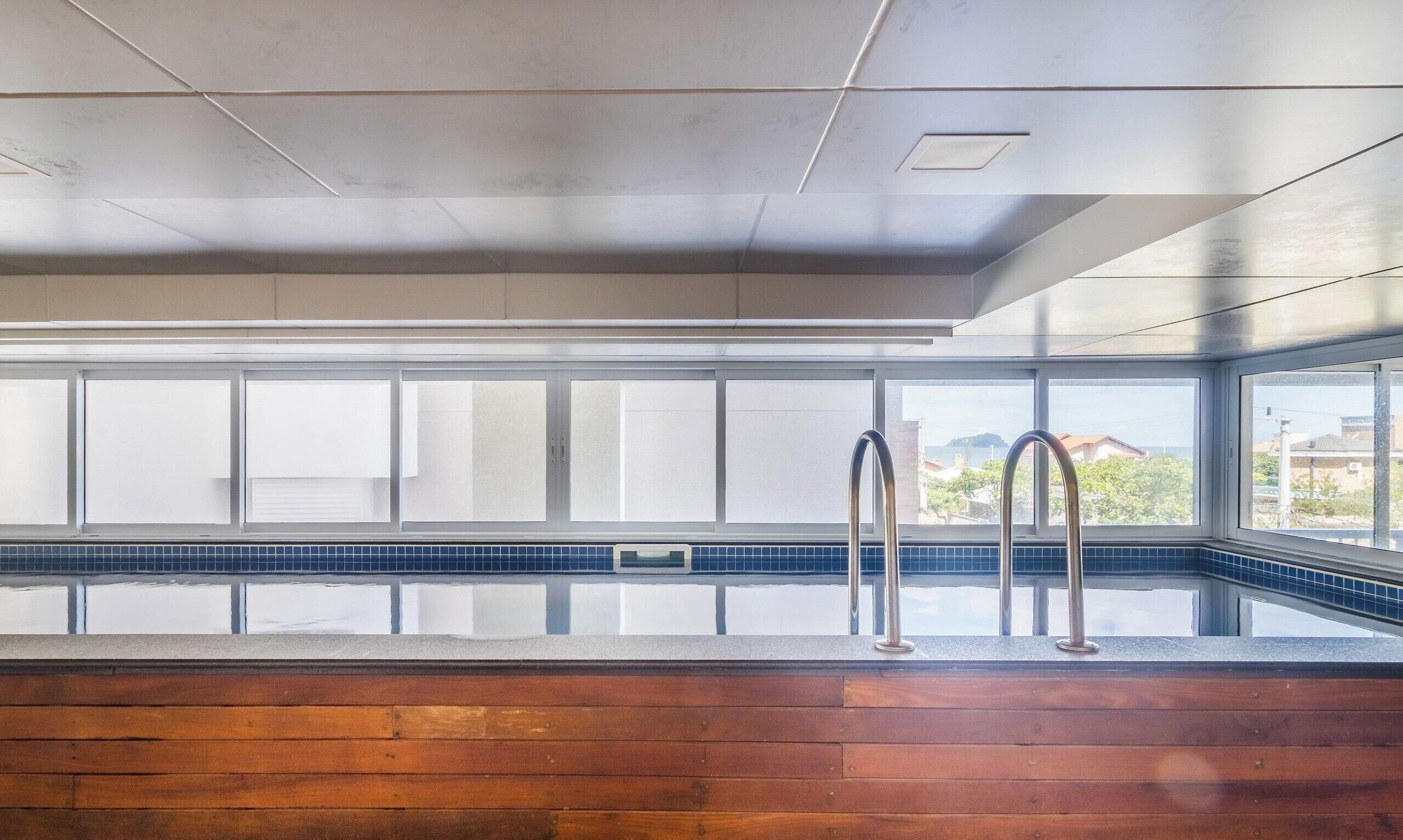 Ein moderner, überdachter Schwimmbereich mit großen Glaswänden, einer Holzterrasse und Metallgeländern.