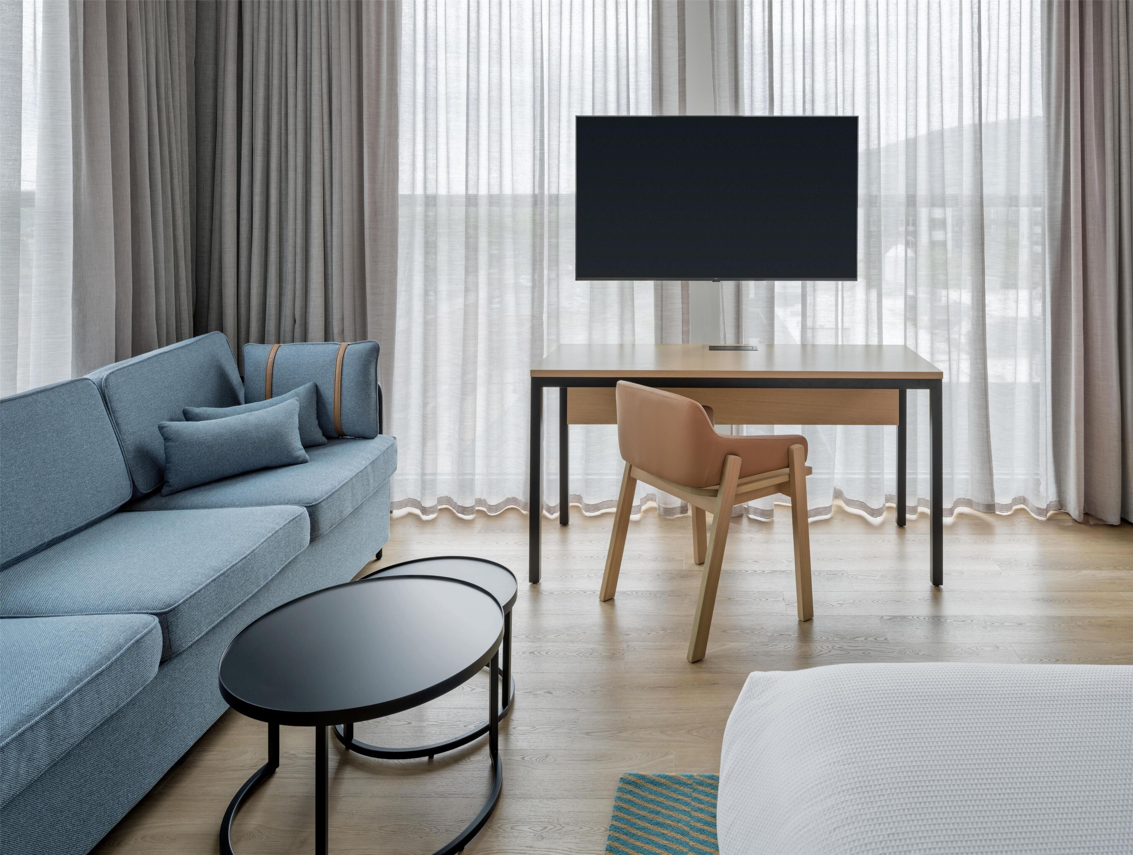Moderni hotellihuone, jossa on sohva, työpöytä, tuoli, televisio ja sänky.