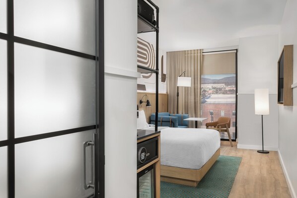 Une chambre d’hôtel avec un lit, un bureau doté d’une lampe, une chaise et une vue sur le paysage urbain, visible à travers une grande fenêtre.