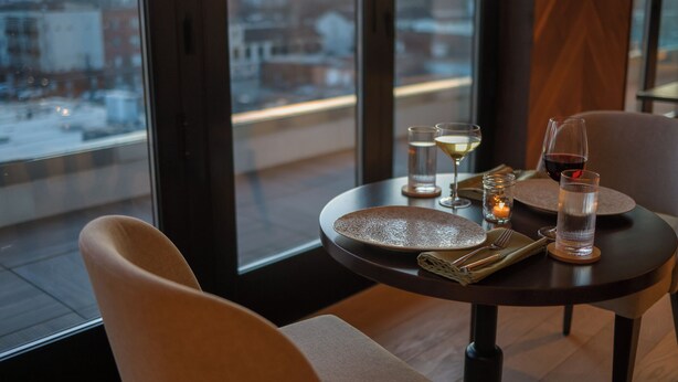 A dining table with plates, glasses, and cutlery, set against a cityscape view through large windows.