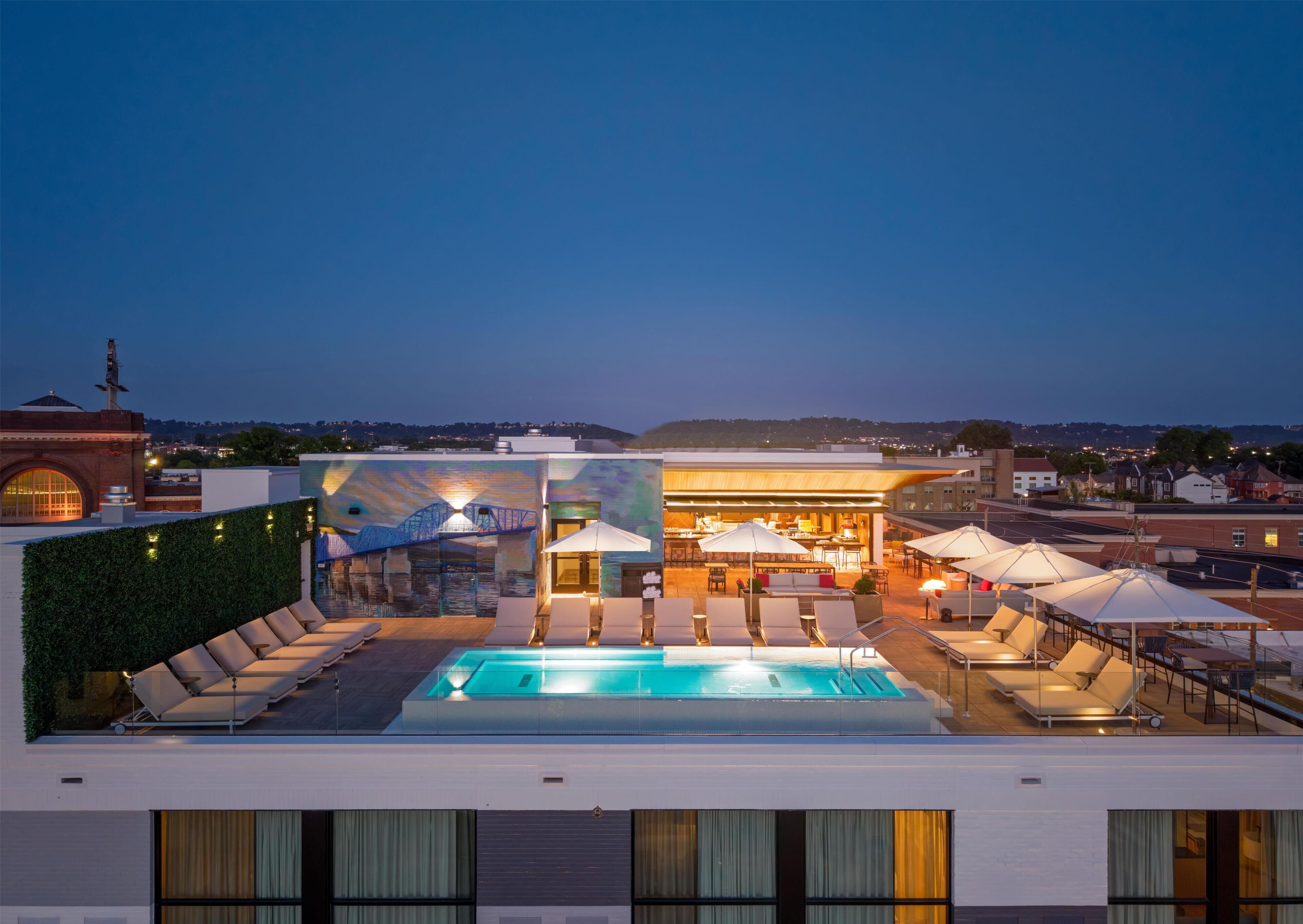 Ein Poolbereich auf dem Dach mit Liegestühlen, Sonnenschirmen, einer Bar und Blick auf die Stadtlandschaft.